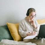 A focused woman in glasses and headphones works on a laptop from a cozy bed.